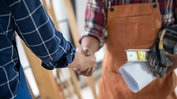 Zwei Personen schütteln sich die Hände, eine Person trägt ein kariertes Hemd und die andere eine Schürze und hält Baupläne sowie Arbeitshandschuhe.