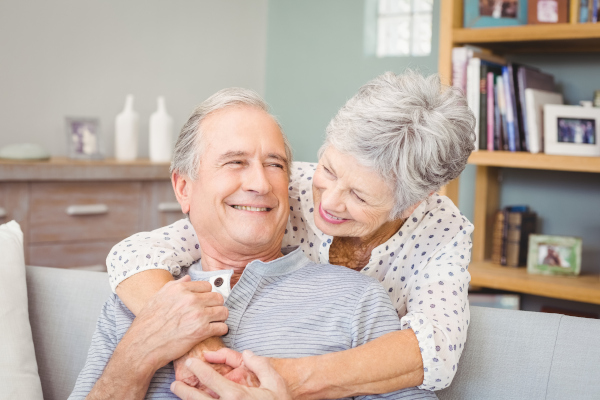 Zwei ältere Personen sitzen auf einem Sofa, wobei eine Person die andere von hinten umarmt.