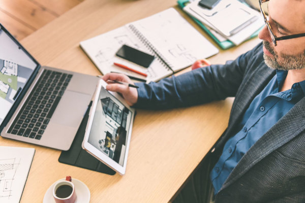 Ein Mann sitzt am Schreibtisch und betrachtet Baupläne sowie ein 3D‑Modell einer Immobilie auf seinem Laptop.